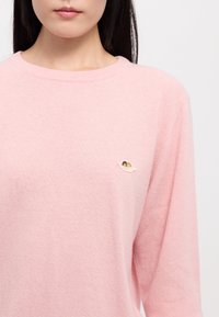 Pull en maille rose clair avec col rond et manches longues. Présente un petit logo coquillage brodé sur le côté gauche de la poitrine. Texture douce.
