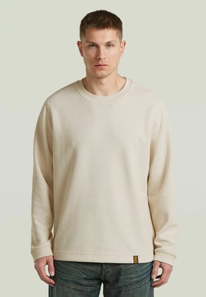 Jonge man met kort haar die een beige trui met lange mouwen en een blauwe spijkerbroek draagt, staand tegen een effen lichte achtergrond.