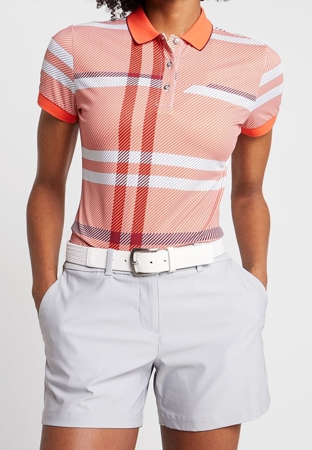 Personne portant un polo à carreaux rouges et blancs avec un col orange, un short gris clair et une ceinture blanche, les mains dans les poches, vue des épaules vers le bas.