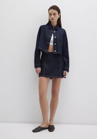 Dark denim cropped jacket and matching skirt, featuring a button front, chest pocket, and contrasting stitching. Model wears a white top and slip-ons.