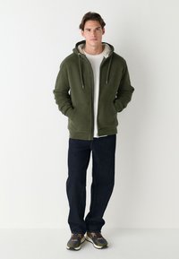 Olijfgroene hoodie met rits en gevoerde capuchon, voorzakken, gecombineerd met donkere spijkerbroek en meerkleurige sneakers op een effen achtergrond.