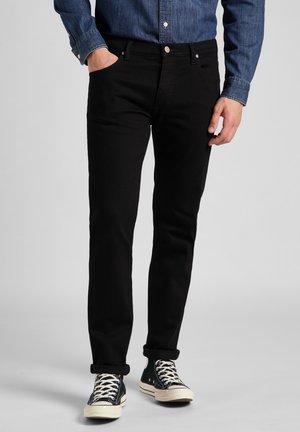 Man die zwarte spijkerbroeken, marineblauwe hoge sneakers met witte veters en een blauw denim overhemd met knopen draagt, staand met één hand in de zak.