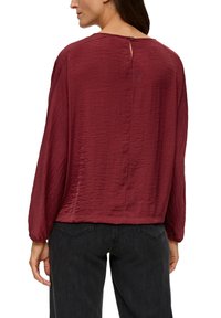 Rote, zerknitterte Bluse aus Stoff mit langen Ärmeln und einem Schlüssellochverschluss hinten, kombiniert mit dunklen Jeans.