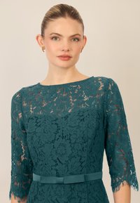 Abito in pizzo teal con maniche a tre quarti, motivi floreali, bordi scallop e una cintura di seta che accentua il punto vita. Gioielli minimalisti in evidenza.