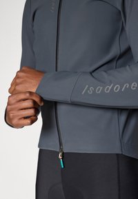 Chaqueta de cremallera en gris oscuro hecha de un tejido suave y elástico con una cremallera negra y un logo sutil en la manga. Diseño ajustado con mangas largas.