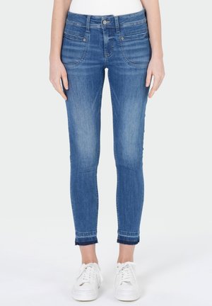 Femme portant un jean skinny bleu taille mi-haute avec poches avant et baskets blanches, debout devant un fond uni.