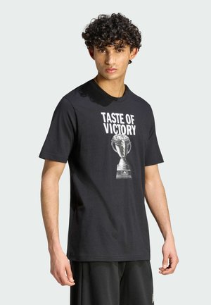 Giovane uomo con capelli ricci indossa una maglietta nera con la scritta "TASTE OF VICTORY" e grafica di un trofeo, in piedi contro uno sfondo chiaro uniforme.