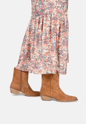 Bottes de cowboy en daim marron portées avec une jupe midi fleurie fluide aux tons pêche, bleu et crème sur un fond uni.