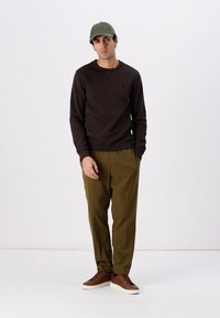 Brauner Pullover mit Rundhalsausschnitt, kombiniert mit olivgrünen Hosen und braunen Schuhen. Das Model trägt eine hellgrüne Mütze. Schlichte, lockere Passform.