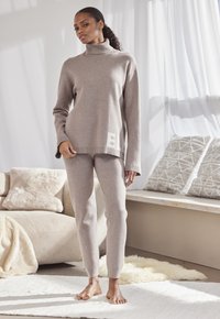 Beige gestrickter Rollkragenpullover mit langen Ärmeln und seitlichen Schlitzen, kombiniert mit passenden schmal geschnittenen Hosen. Verfügt über ein dezentes Logodetail.