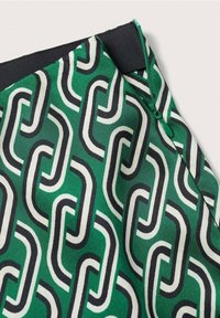 Tissu à motif géométrique de maillons de chaîne verts, noirs et blancs, avec une ceinture élastique noire et une fermeture éclair verte sur un vêtement.