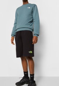 Modrý svetr s kulatým výstřihem a logem, kombinovaný s černými šortkami s zelenými logovými akcenty. Obě části jsou vyrobeny z měkké látky.