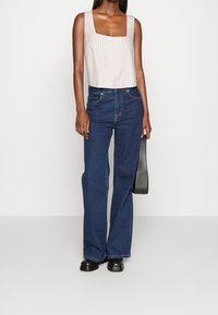 Beige randig kort tanktopp med fyrkantig halsringning, kombinerad med mörkblå flarejeans och svarta skor, håller en svart väska.