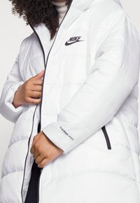 Biała pikowana kurtka puchowa z błyszczącego materiału, z przednim zamkiem błyskawicznym, kapturem i czarnym logo Nike. Napis "THERMAFIT" na mankiecie. Pikowany wzór.