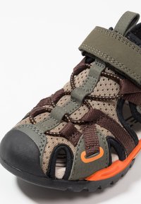 Sportssandal med en andningsbar beige mesh-överdel, mörkbruna remmar, gröna accenter och en orange sula med en robust svart yttersula.