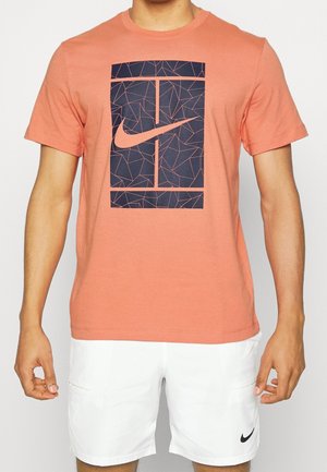Mężczyzna w koralowej koszulce z granatowym geometrycznym logo Nike i białych szortach z małym czarnym logo Nike.