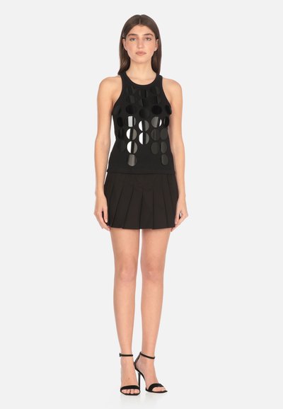 Top nero senza maniche con ritagli circolari, abbinato a una gonna nera plissettata e sandali neri con tacco alto. Tessuto liscio, design aderente.