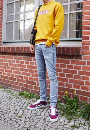 Geel crewneck sweatshirt met logo, lichtblauwe skinny jeans en kastanjebruine sneakers met witte accenten. Gevoelige luipaardprint tas.