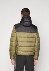 Giacca imbottita con corpo verde oliva e area delle spalle nera. Presenta un cappuccio e una texture a righe orizzontali. Finitura opaca, senza hardware visibile.