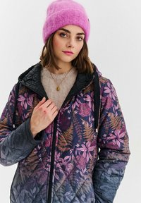 Mladá žena v růžové pletené čepici, květinově vzorované prošívané bundě s černou kapucí, béžovém svetr a zlatém přívěsku, se dívá do kamery.