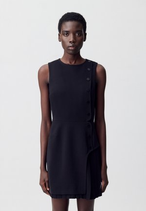 Una donna con capelli corti indossa un vestito nero senza maniche con dettagli di bottoni e un orlo in plissé, posando su uno sfondo neutro.
