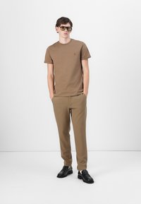 T-shirt marron à manches courtes avec un petit logo, associé à un pantalon fuselé assorti et des chaussures noires à enfiler ; design et texture minimalistes.