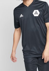 Camiseta atlética negra de mangas cortas, con un diseño de cuello en V. Logo blanco de Adidas y un gráfico hexagonal en el pecho.