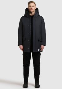 Czarna izolowana kurtka z kapturem, z zapięciem na guziki, dwoma przednimi kieszeniami i lekko pikowanym wzorem, noszona na czarnych spodniach i butach.