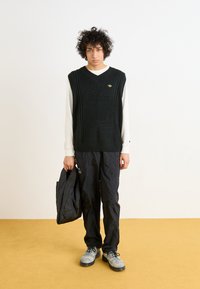 Pull en tricot vert sans manches avec un col en V, porté par-dessus une chemise blanche à manches longues ; associé à un pantalon noir et des chaussures grises, tenant une veste noire.