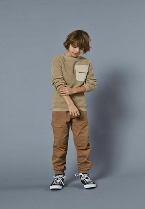Tan strikket sweater med en cremefarvet lomme, parret med brune cargo bukser og sorte sneakers med hvide detaljer. Enkelt, afslappet outfit.