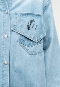 Ανοιχτό μπλε τζιν πουκάμισο με ασύμμετρη εμπρόσθια τσέπη που φέρει το κείμενο "MOSCHINO 86-23 JEANS" και ασημένια κουμπιά πίεσης στο εμπρόσθιο μέρος.