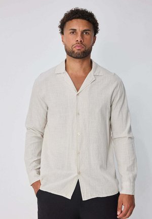 Homme aux cheveux courts et bouclés avec une barbe, portant une chemise crème à manches longues boutonnée et un pantalon noir, debout devant un fond uni.