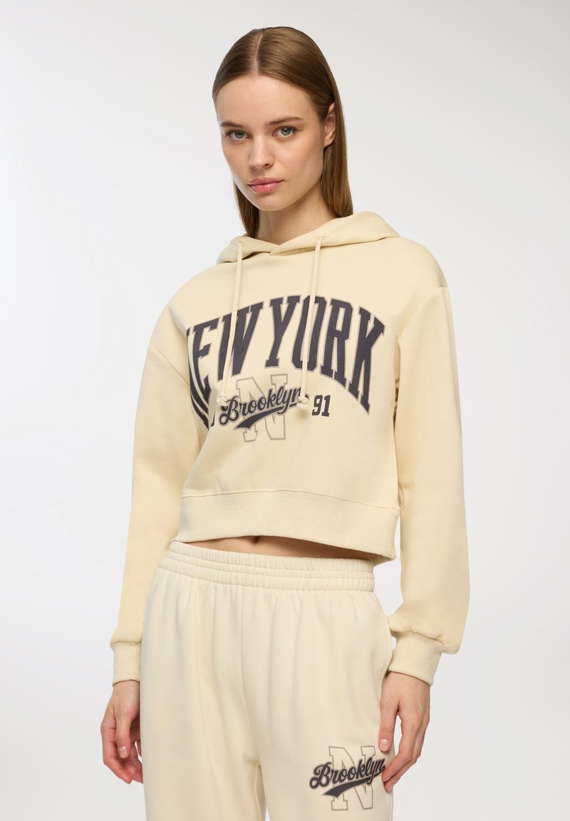 Kremowy, krótki hoodie z nadrukiem "NEW YORK" i "Brooklyn" w ciemnoszarym kolorze, z kapturem i sznurkami; dopasowane spodnie dresowe.