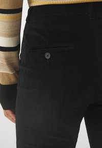 Pantalon en velours côtelé noir avec une poche arrière ornée d'un bouton noir rond. Le tissu présente une texture côtelée. Un pull rayé est partiellement visible.