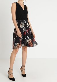 Robe noire sans manches avec un décolleté en v croisé, ornée d'une jupe à motifs floraux dans des nuances de rouge, blanc et jaune ; accompagnée de talons noirs à lanières.