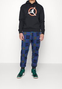 Sudadera negra con un logo en blanco y naranja, combinada con pantalones de cuadros azules que presentan acentos en verde y rojo, completados con zapatillas deportivas verdes de caña alta.