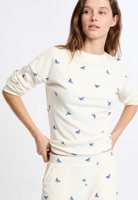 Mujer ajustándose una blusa de manga larga color crema y pantalones con pequeños patrones de lazo azul bordados sobre un fondo blanco.