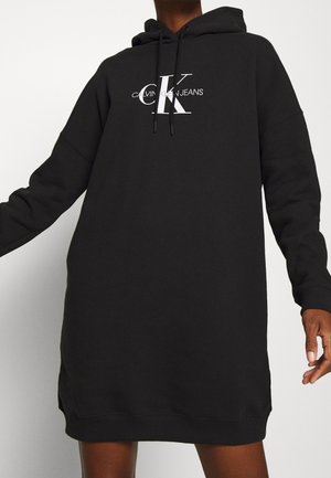 Osoba ubrana w czarną długą sukienkę z kapturem z logo "Calvin Klein Jeans" w białym kolorze na piersi oraz regulowanymi trokami kaptura.