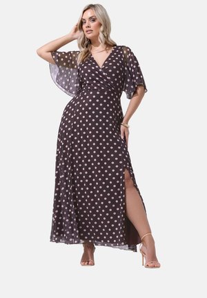 Femme portant une robe maxi noire à pois rose pâle, manches flottantes, col en V, fente latérale, et sandales à talons beiges, posant avec la main sur la tête.