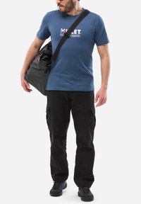 Niebieska bawełniana koszulka z białym nadrukiem graficznym, czarne spodnie cargo, czarne buty oraz szara torba na ramię z paskiem.