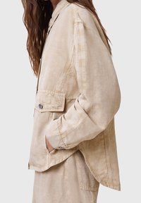 Femme aux longs cheveux bruns portant une veste beige oversize et un pantalon large assorti, main dans la poche de la veste, de profil.