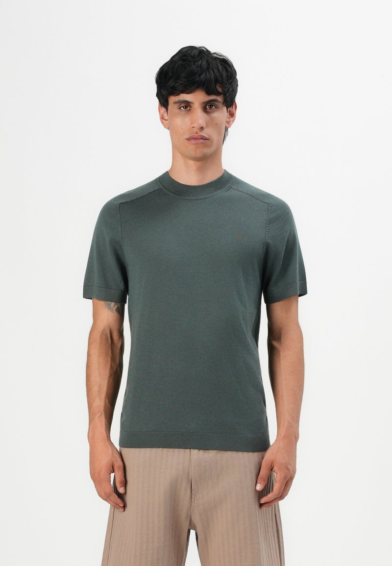 Homem de pé contra um fundo simples, vestindo uma camiseta de malha de manga curta verde-escura e calças plissadas beige claro.