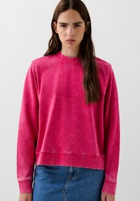 Felpa fucsia a girocollo con polsini e orlo a coste, caratterizzata da una vestibilità morbida e una texture leggermente sbiadita. Abbinata a una gonna di jeans.