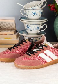 Paire de baskets roses pailletées avec des bandes blanches, tasses à thé vintage empilées avec des motifs floraux bleus, et une pile de livres à proximité.