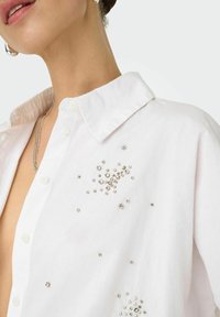 Camicia bianca con bottoni, caratterizzata da un colletto classico e decorazioni in cristallo disposte a grappolo. Tessuto morbido con una vestibilità strutturata.