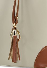 Sac tote avec un corps crème, des accents en cuir marron, une ferrure anneau doré, et un gland fait de lanières de cuir marron et métallisé.