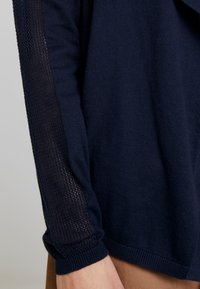 Granatowy sweter robiony na drutach z teksturowanym, otwartym splotem na rękawie. Posiada dopasowane mankiety i gładką część główną, z klasycznym designem okrągłego dekoltu.