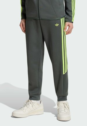 Donkergrijze Adidas trainingsbroek met neongele strepen aan de zijkanten, gecombineerd met witte sneakers, gedragen door een staand persoon.
