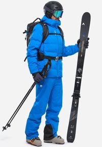 Blaue isolierte Skijacke, passende Hose, schwarzer Helm und Skibrille. Hält schwarze Skier und einen Skistock, trägt einen Rucksack.