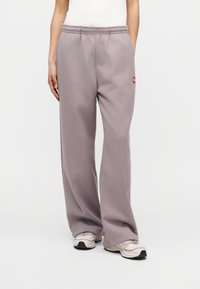 Γυναίκα που φοράει χαλαρό μωβ φόρμα με ελαστική μέση και μπεζ αθλητικά παπούτσια, στέκεται μπροστά σε απλό φωτεινό φόντο.